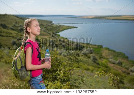 Girl with a backpack standing on the background of the lake and drinking water, sunny day