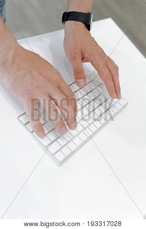 Typing and testing. Scaled up shot of adult male hands with a wrist watch typing on a wireless computer keyboard lying on a white writing table.