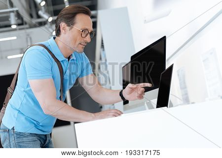 Wait what is that. Adult shopper wearing casual attire leaning on a display and pointing his finger at a template laptop while walking around an electronics shop.