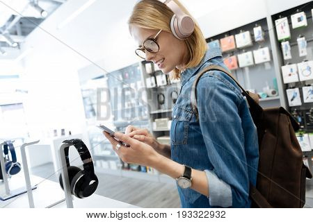   Focused blonde lady turning on the music on her smartphone while testing a pair of stylish mockup headphones at an electronics store.