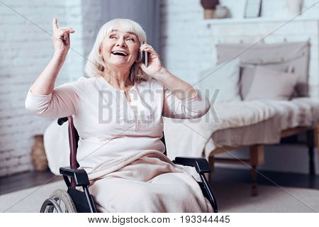 Full of joy. Merry old enable woman sitting in the wheelchair at home while smiling and using mobile