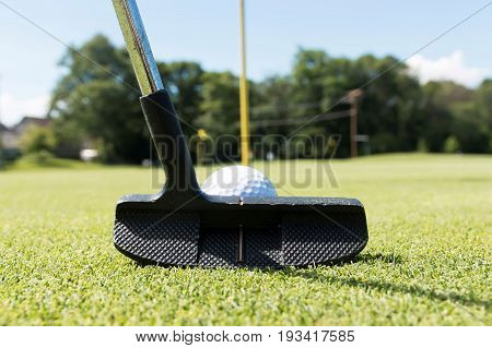 Close up view of a golf putting club with a white golf ball in front of it lining up a put toward the yellow flag pin.