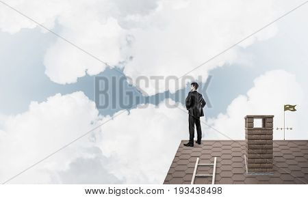 Young determined businessman standing with back on house roof and looking away. Mixed media