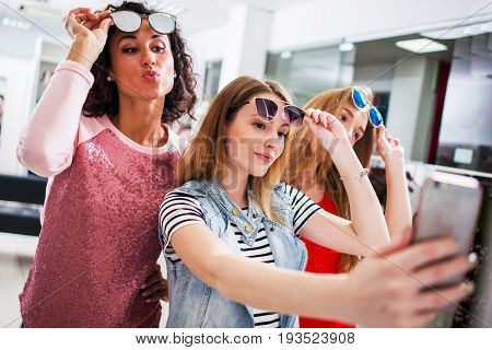 Three young stylish girlfriends raising fashionable sunglasses while taking selfie with smartphone in shopping mall.