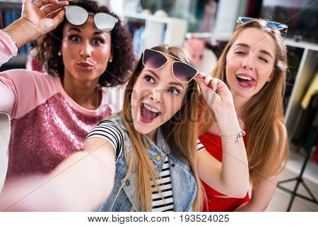 Three female friends taking selfie making faces raising sunglasses in clothing and accessories outlet.