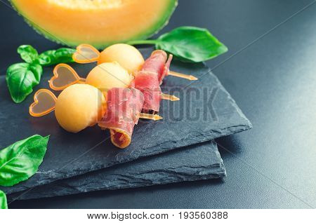 Colorful canape on skewers with melon and prosciutto on a slate plate on dark background. Tasty appetizer for a party or banquet. Concept of italian food. Traditional appetizer antipasto. Copy space.