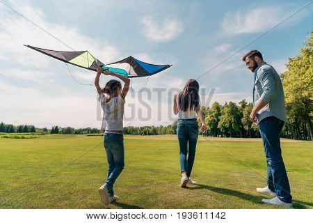 Back View Of Girl Holding Kite With Parents Walking Near By In Meadow