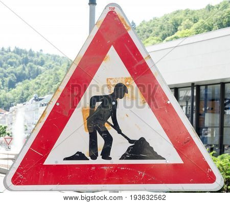 A rusty triangular sign warning of roadworks ahead with a modern city building in the background