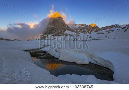 Evening scene in mountains. Sunlit peak reflects in open water. Snow covered Edigol table-land (Aladaglar).