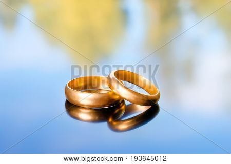 Two bright golden wedding rings on abstract nature background. Reflection with blurred blue sky and green trees