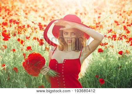 girl with long curly hair in red dress and retro hat hold flower bouquet in field of poppy seed on green stem on natural background summer drug and love intoxication opium pin up woman