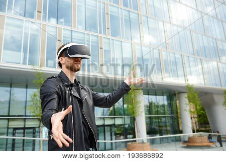 Amazed young Caucasian male in black clothing experiencing virtual reality using oculus rift headset for entertaining himself gesturing as if touching something while playing video game in cityscape