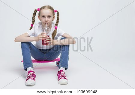 Kids Concepts. Little Caucasian Blond Girl Posing with Pink Pennyboard and Drinking Red Juice from Long Cup Using Straw. Against White. Horizontal Image