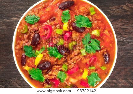 A closeup of chili con carne, a traditional Mexican dish with red beans, cilantro leaves, ground meat, and chili peppers, on a dark rustic texture