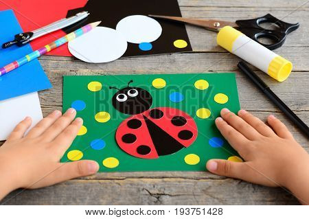 Small child shows a ladybird card. Child made a card with ladybird. Stationery set on a desk. Preschool paper project. Developing children fine motor skills