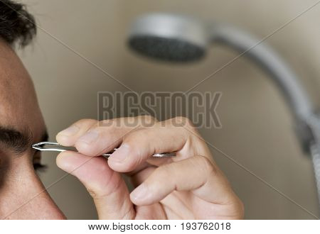closeup of a young caucasian man in the bathroom plucking his eyebrows with tweezers