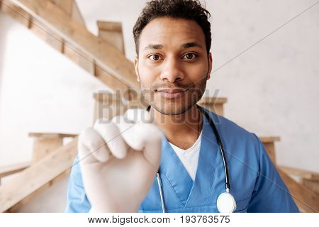 Open your mouth. Delighted international male person wrinkling his forehead and keeping stethoscope on his neck while holding his hand on the foreground