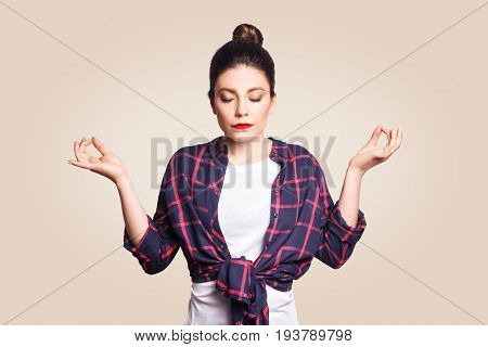 Meditation religion and spiritual practises. Beautiful young brunette girl doing yoga in the morning indoors at beige wall after waking up keeping eyes closed holding fingers in mudra gesture