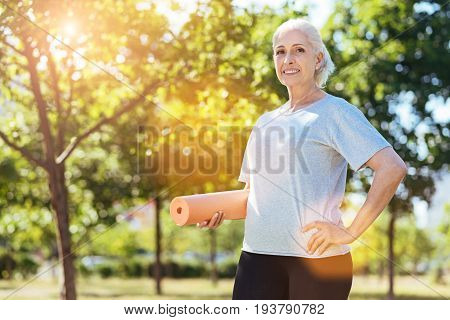 Full of gladness. Cheerful aged woman holding joga carpet and smiling while doing exercises in the park