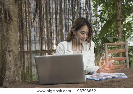 Asian smile beautiful girl sitting outdoor office using smart phone and laptop in nature. Work space with work sheet of business business and lifestyle conceptcopy space