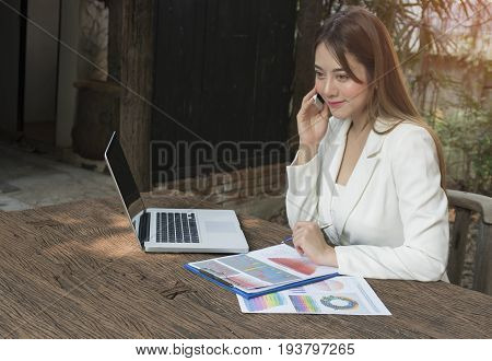 Asian smile beautiful girl sitting outdoor office using smart phone and laptop in nature. Work space with work sheet of business business and lifestyle conceptcopy space
