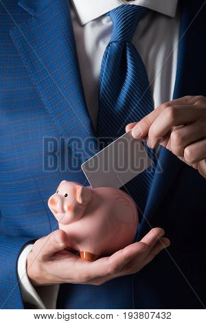 Banking money loan moneybox ecash and information. Male hands putting blank business card in piggy bank for saving on grey background. Fashionable formal suit and tie