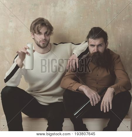 health and addictive. Young man with milk or yogurt and bearded hipster with wine. Two serious friends in casual wear sitting with bottles on beige wall. Friendship convive bad habits