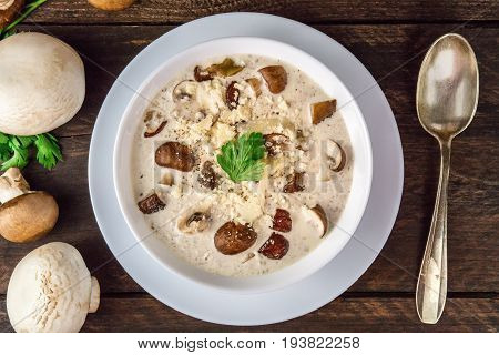 An overhead photo of a plate of a mushroom cream soup with champignons, cremini, fresh parsley, and a spoon, with grated cheese, on a dark rustic background texture