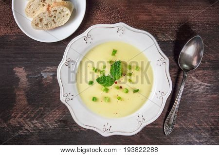 Overhead photo of vichyssoise, French potato and leeks soup, with chopped green onions, pink peppercorns, and mint leaves, on dark rustic texture with bread, spoon, and place for text