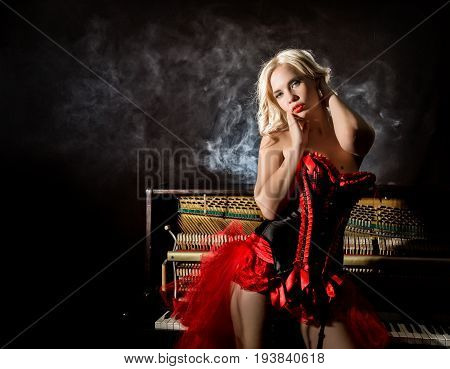 beautiful blonde musician in red corset standing near the piano. Vintage style beautiful woman.. Old fashioned makeup and retro finger wave hairstyle.