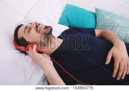 Attractive young man listening to music and relaxed in bed. Indoors.