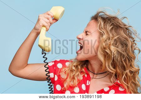 Blonde curly haired woman wearing red dress with white dots yelling at phone receiver standing against blue background.