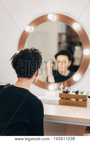 Attractive woman primp at the mirror with lamps in the beauty studio. A happy satisfied customer of make-up and hairstyle service.