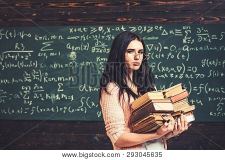 Brunette Student Walking With Two Heaps Of Books In Front Of Green Board Full Of Writing. Math Under