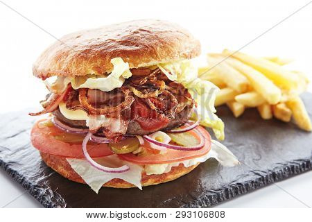 Fresh Beef Burger with Bacon, Fried Onions, Tomato Sauce, Pickled Cucumbers, Green Lettuce, Cheese and French Fries Garnish. Hamburger or Beefburger on Natural Black Stone Plate Isolated on White