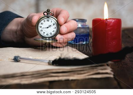 Man Is Holding In Hand A Pocket Watch On A Brown Table Background With A Documents And Burning Candl