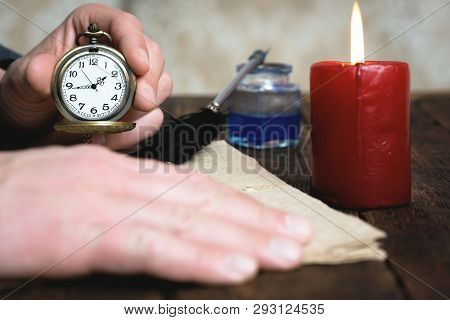 Man Is Holding In Hand A Pocket Watch On A Brown Table Background With A Documents And Burning Candl