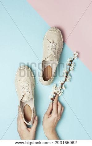 Female Hands Hold Beige Fashionable Espadrilles With Spring Flowering Branch On Pastel Blue Pink Bac