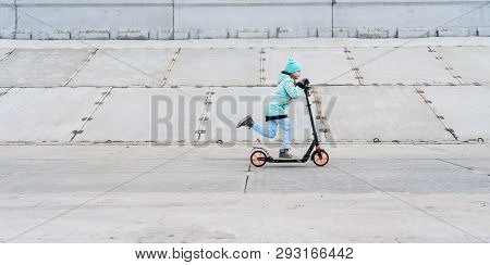 Activities: A School Girl With A Blue Jacket Riding A Scooter On A Gray Concrete Promenade.