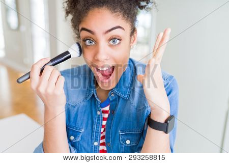 Young african american girl appying blush using make up brush very happy and excited, winner expression celebrating victory screaming with big smile and raised hands