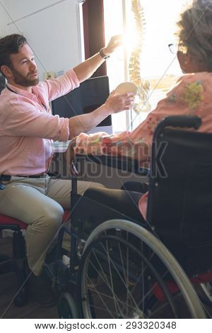 Side view of confident Caucasian male doctor discussing over anatomical model with senior woman in clinic 