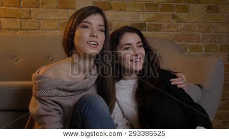 Closeup Portrait Of Two Young Pretty Women Hugging And Watching Tv With Excitement In A Cozy Apartme