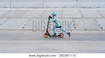 Activities: A School Girl With A Blue Jacket Riding A Scooter On A Gray Concrete Promenade.