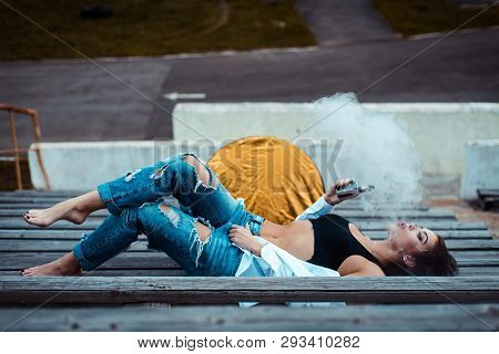 Young Sexy Woman Lying On The Floor And Smoking Vaping E-cigarette. She's Wearing A Top And A White 