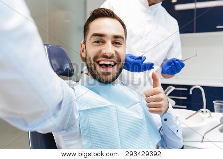 Image of a handsome happy young man sitting in medical dentist center take a selfie by camera make thumbs up.