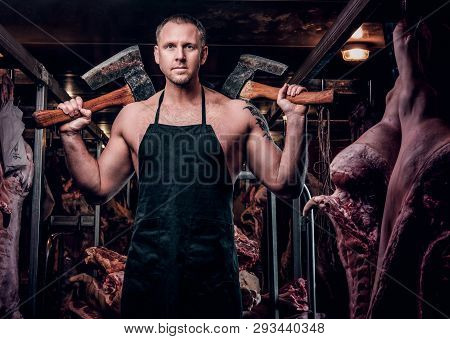 Shirtless Butcher With A Tattoo On His Hand Wearing Apron Posing With Two Axes In A Refrigerated War