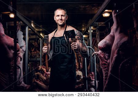 Smiling Butcher With A Tattoo On His Hand Wearing Apron Posing With Two Axes In A Refrigerated Wareh