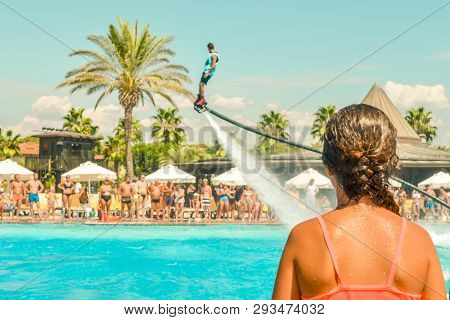 Teen Girl Watching Fly Board Watershow At The Pool. View From The Back. Summer Holidays, Fun Summert