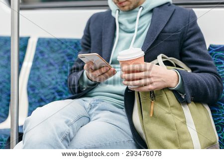 Cropped Photo Of Handsome Man In Blue Coat And Turquoise Hoodie Is Sitting In A Subway Train, Drinki