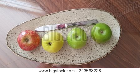 A Plate With Four Apples Of Various Colors And A Knife Stay On Glass Table In Sunlight